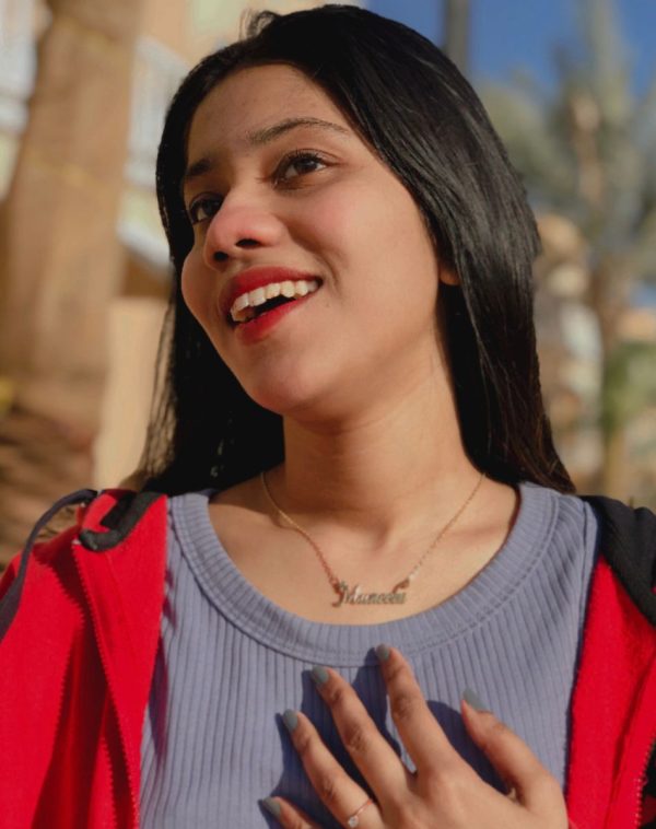 Muneeba wearing Single Name Gold-Plated Necklace, laughing and pointing to her name, grey ribbed sweatshirt with open red hoodie.