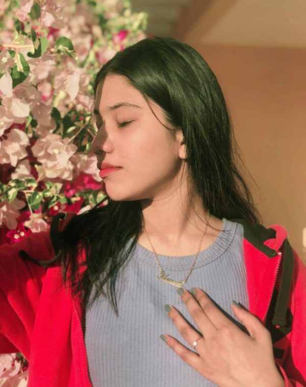 Muneeba with closed eyes, smelling flowers, wearing green matte nail polish and a ring, Single Name Gold-Plated Necklace.
