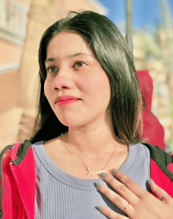 Muneeba pointing to her name, wearing grey ribbed sweatshirt with open red hoodie, Single Name Gold-Plated Necklace.