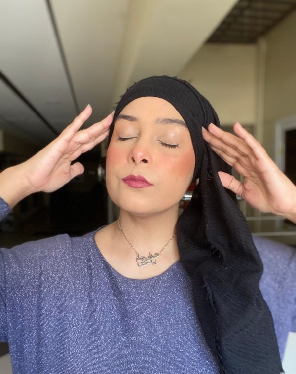 Influencer Khadija Yaseen showcases our Double Arabic Name Silver Plated Necklace, wearing a blue top and a black scarf, with her hands on her head and eyes closed in a captivating pose.