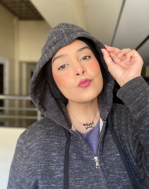 Influencer Khadija Yaseen radiates charm with our Double Arabic Name Silver Plated Necklace, wearing a grey hoodie, striking a Korean heart pose in a captivating selfie.
