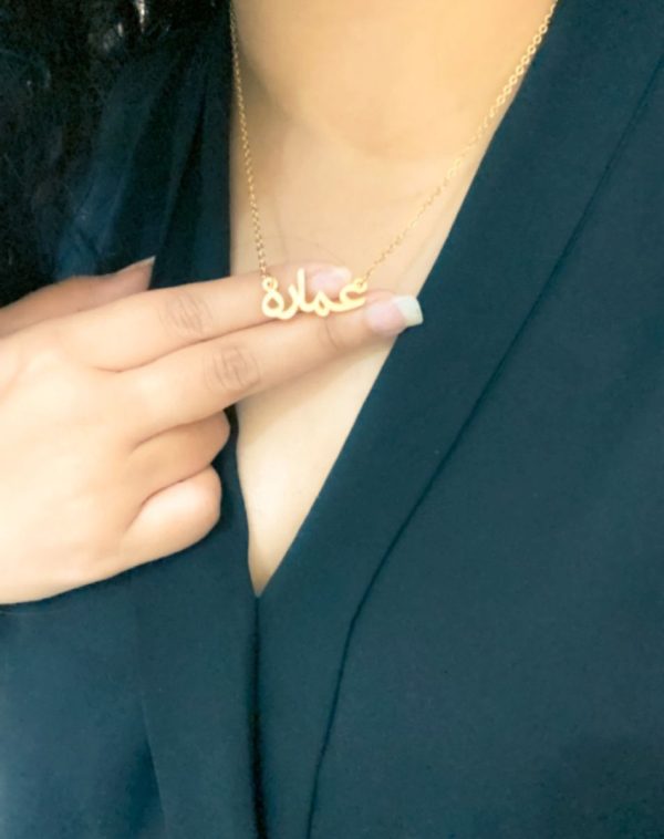 Close-up shot of the Single Arabic Name Gold-Plated Necklace with the customized name pendant "Ammara" held between two fingers, visible from the chin to the chest of the influencer Ammara.