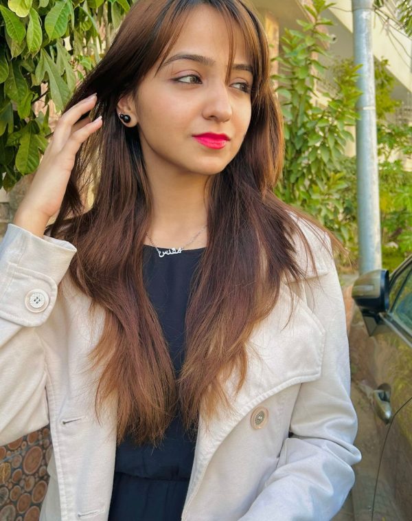 Influencer Muqaddas showcases our Single Arabic Name Silver-Plated Necklace in a captivating outdoor photo. She wears an off-white fleece jacket with a black inner, posing with hands on hair between shoulders and ear, looking away from the camera.