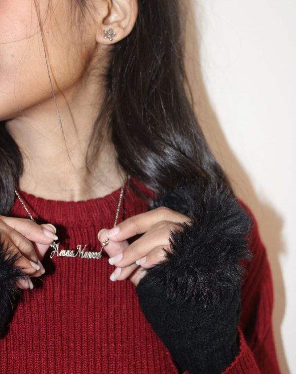 Close-up of Amna Masood wearing a Double Name Silver-Plated Necklace, her face partially visible with a red sweater and black fur-edged gloves, holding the necklace with both hands.