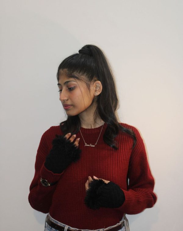 Full-length portrait of Amna Masood wearing a blood red ribbed sweater, black gloves with fur edges, and the Double Name Silver-Plated Necklace. She stands against a plain white wall, posing with hands on chest and stomach.