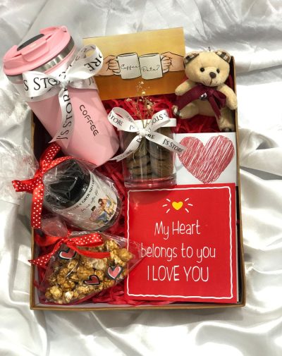 The Coffee Date Gift Box For Her is a Valentine's Day gift package that includes a pink temperature indicating thermal coffee mug, a coffee jar for storing coffee grounds or beans, a cookie jar filled with chocolate chip cookies, a pack of Salted Caramel popcorn, and a cute stuffed teddy bear. The gift box also includes a Valentine's Day special greeting card and is beautifully packed in a box packaging, making it ready to gift.