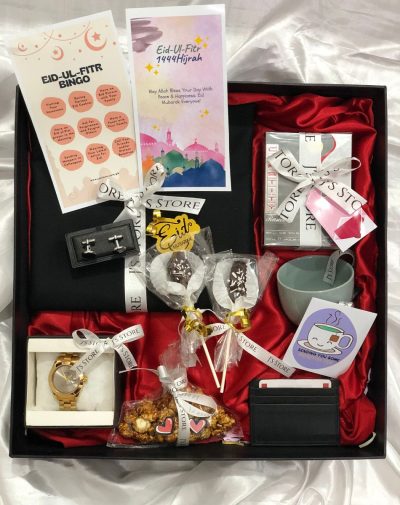 A curated gift box with various items for Eid celebrations, including an unstitched Shalwar Kameez suit, perfume, watch, marble cufflinks, a leather cardholder, two chocolate coated date pops, a bag of salted caramel popcorn, a teacup with tea bags, an Eid bingo game, and an Eid greeting card. The Festive Eid Box comes in a 16 * 16 inches curated box with Imported Shredded Papers, ribbons, mini cards, and more, making it a beautiful and thoughtful gift.