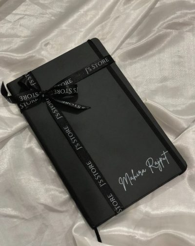 An open shot of the Personalized Leather Notebook - Black with a customized name on the cover in silver, emphasizing the creative element. The notebook is decorated with Black J's Store's ribbon.