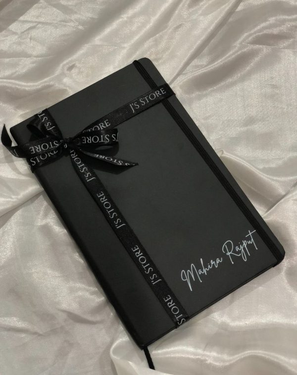 An open shot of the Personalized Leather Notebook - Black with a customized name on the cover in silver, emphasizing the creative element. The notebook is decorated with Black J's Store's ribbon.