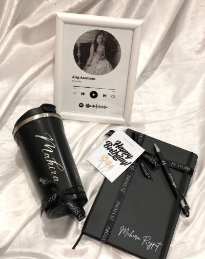 A curated gift box showcasing a white Spotify frame playing a song with spiral artwork, a black temperature-indicating coffee mug with a name engraved, a black leather notebook with silver name personalization, and an engraved metal pen, all arranged on a white surface.