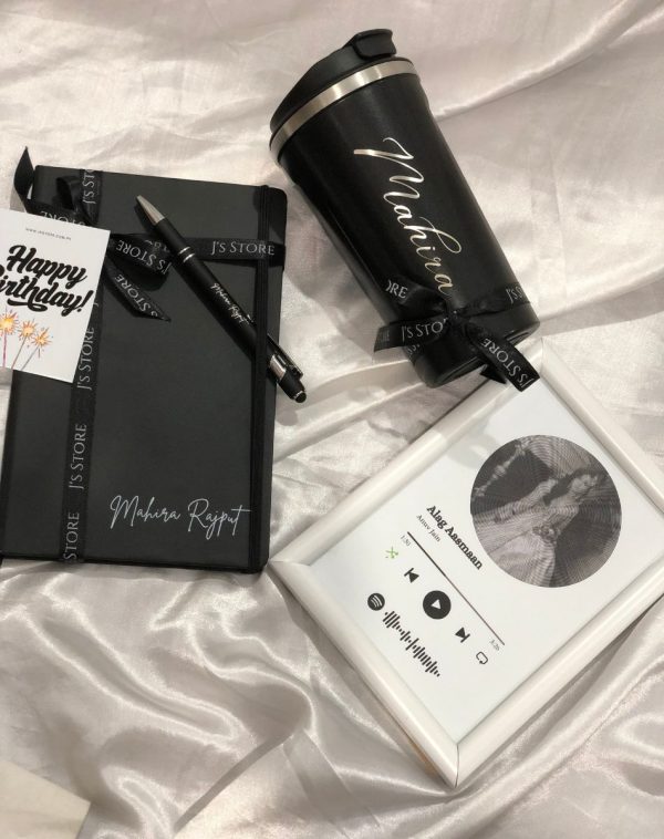 A curated selection of personalized treats spills from a beige gift box: a white Spotify frame displaying a song with spiral betty artwork, alongside a black temperature-indicating coffee mug with a name engraved, a black leather notebook with silver name personalization, and an engraved metal pen, all resting on a textured surface.