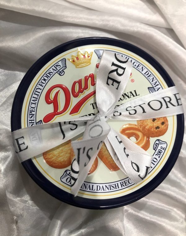 Danisa Cookies: A round, blue and white tin of Danisa Traditional Butter Cookies. The lid features the red "Danisa" logo with a crown, images of butter cookies, and text indicating "Traditional Butter Cookies" and "Original Danish Recipe." A white ribbon with black text that includes "J's STORE" is tied around the tin.