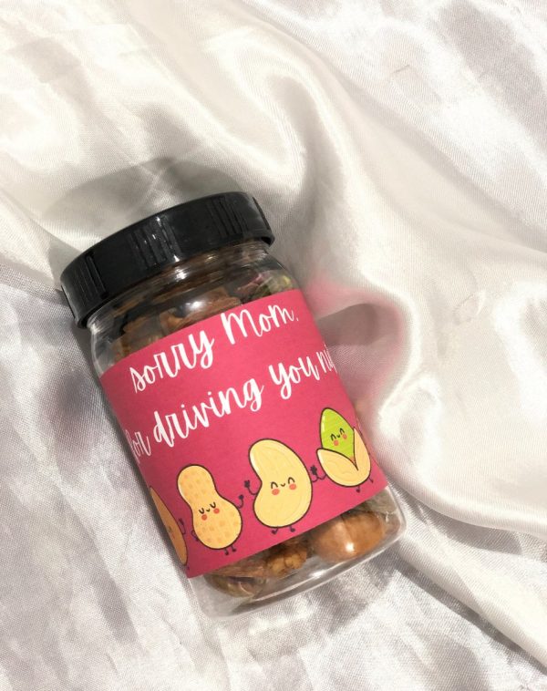 Dry Fruits Jar For Mother's Day: A clear plastic jar with a black lid, filled with a mix of dry fruits. A pink label wraps around the jar with the handwritten text "Sorry Mom for driving you nuts!" in white. Below the text are cartoon illustrations of peanuts and a pistachio with cute faces.