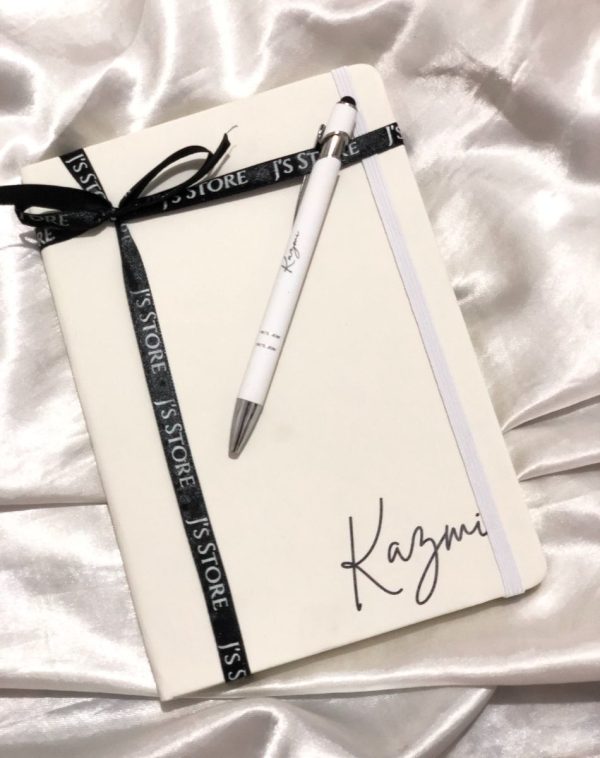 A white leather notebook and a matching pen. The notebook is personalized with the name 'Kazmi' and tied with a black ribbon.