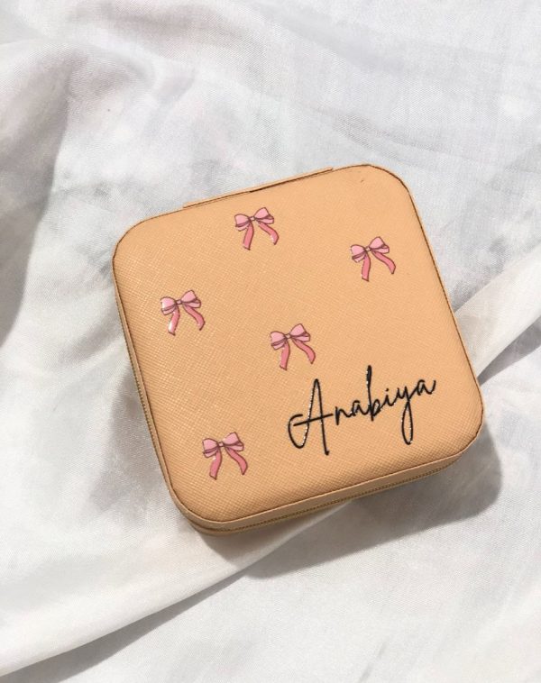 Personalized pink (or nude/black/white) jewelry travel case with pink bow decals and a custom name ("Sania," "Maha," "Fiza," or "Anabiya") in script font, tied with a ribbon.