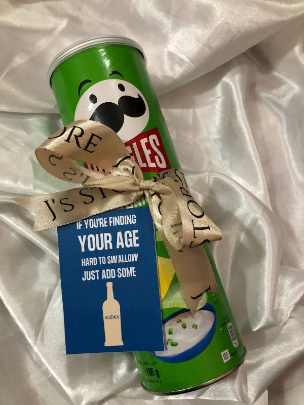 A green Pringles Sour Cream & Onion can wrapped in a beige "J's Store" branded ribbon with a decorative bow. Attached is a blue gift tag that reads, "If you're finding your age hard to swallow just add some vodka," featuring a white silhouette of a vodka bottle. The gift is resting on a ruffled white satin fabric background.