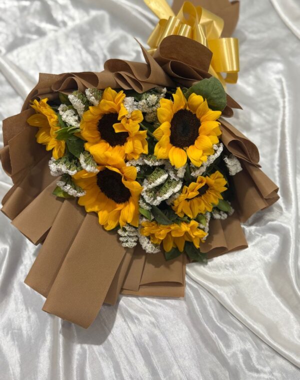Autumn Glow Sunflower Bouquet featuring fresh yellow sunflowers and white fillers in brown wrapping with a gold bow displayed flat on white silk at J’s Store